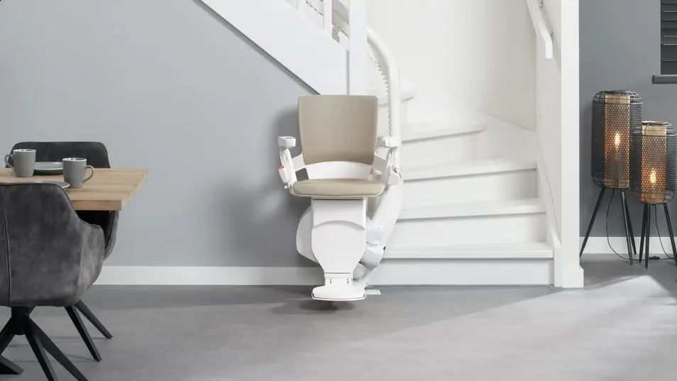 Moderner Treppenlift mit sandfarbenem Polster am Fuß einer weißen kurvigen Treppe