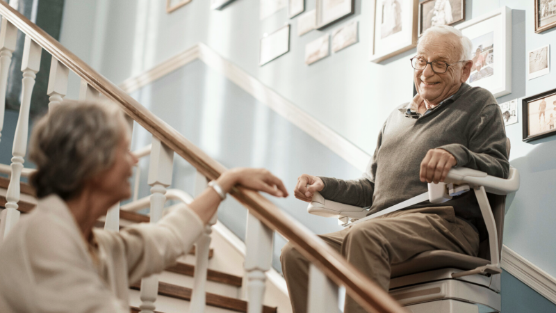 Ein älterer Mann fährt mit einem Sitzlift die Treppe hinauf und lächelt dabei seine Ehepartnerin an