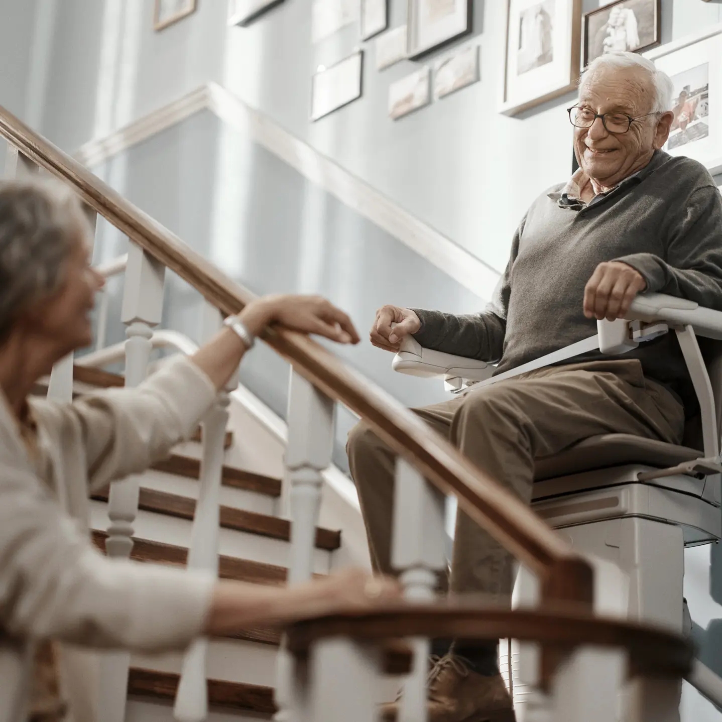 Ein älterer Mann fährt mit einem Sitzlift die Treppe hinauf und lächelt dabei seine Ehepartnerin an