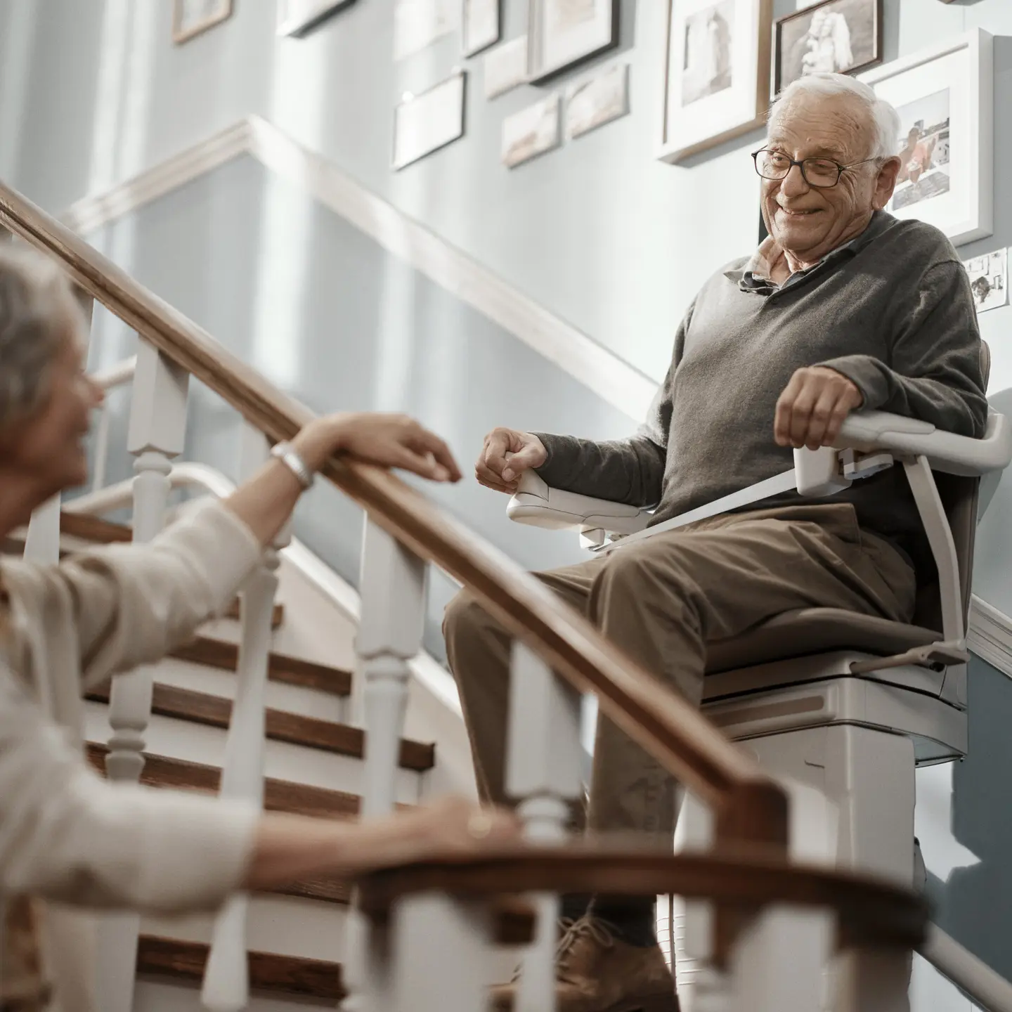 mann_im_lift_neu.jpg Ein älterer Mann fährt mit einem Sitzlift die Treppe hinauf und lächelt dabei seine Ehepartnerin an