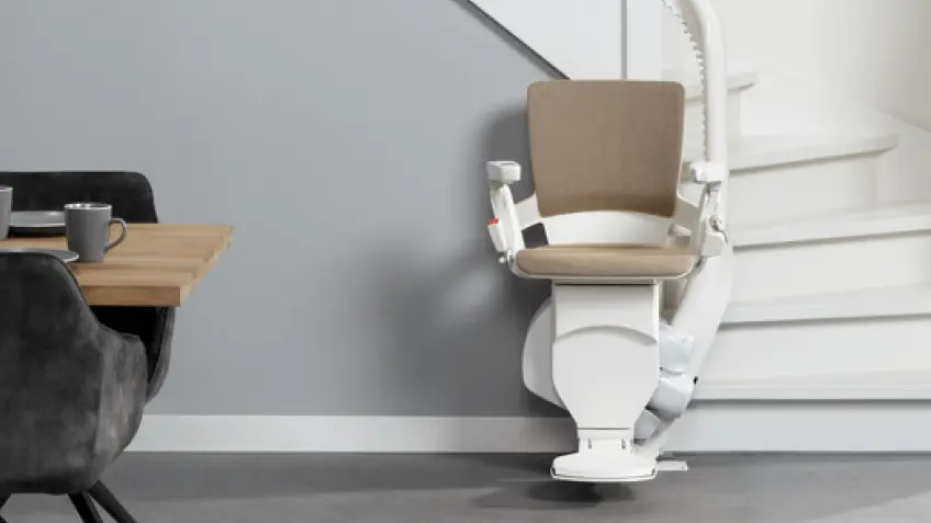 markenteaser-applus.png Moderner Treppenlift mit sandfarbenem Polster am Fuß einer weißen kurvigen Treppe